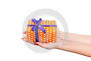 Woman hands give wrapped Christmas or other holiday handmade present in orange paper with purple ribbon. Isolated on white