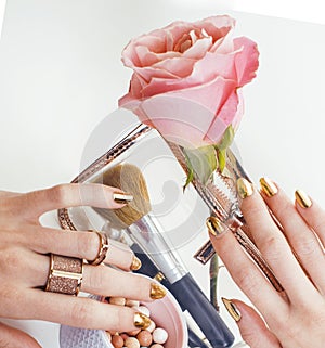 woman hands with golden manicure and many rings holding brushes, cosmetic and rose flower on white background, spa