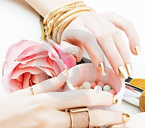 woman hands with golden manicure and many rings holding brushes, cosmetic and rose flower on white background, spa