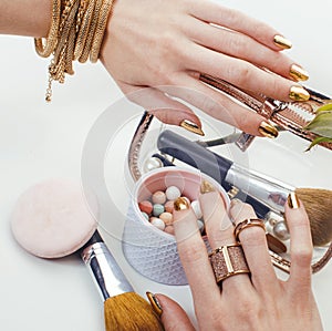 woman hands with golden manicure and many rings holding brushes, cosmetic and rose flower on white background, spa