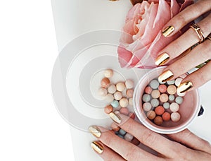 woman hands with golden manicure and many rings holding brushes, cosmetic and rose flower on white background, spa