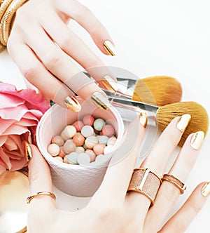 woman hands with golden manicure and many rings holding brushes, cosmetic and rose flower on white background, spa