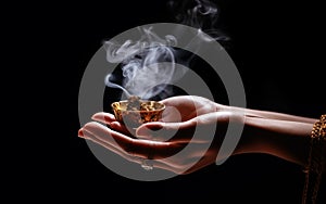 woman hands hold an ornamental incense from which smoke rises, black background.