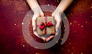 Woman hands holding a Christmas gift box decorated with a red ribbon and golden glitter on red background. AI generated