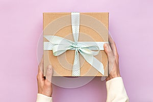 Woman hands holding craft gift box with blue bow on pastel pink background. Girl binds classic bow for present box. Top view, flat