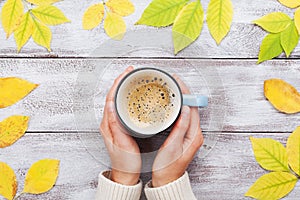 Woman hands holding morning cup of coffee on vintage wooden table decorated autumn yellow leaves top view. Cozy fall breakfast.