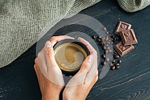 Woman hands holding mug of hot coffee, standing on wooden table with chocolate, coffee beans and sackcloth