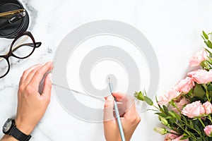 Woman hands holding pencil and write on blank paper card mockup over modern home office desk workspace with stationery, rose