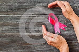 Woman hands holding pink ribbon on wooden background. Top view, with copy space for text. Breast cancer awareness.