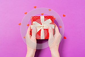 Woman hands holding Red gift present box with small red heart around on purple background. Mother`s Day concept. St