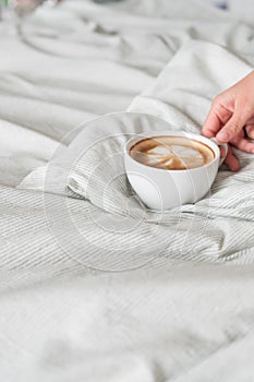 Woman hands holds coffee in white cup on grey sown bed linen. Latte art on top view. Space for text