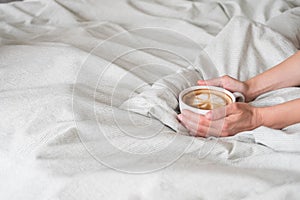 Woman hands holds coffee in white cup on grey sown bed linen. Latte art on top view. Space for text