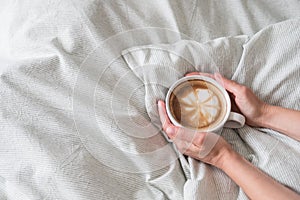 Woman hands holds coffee in white cup on grey sown bed linen. Latte art on top view. Space for text