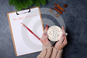 Woman hands holds mug with cacao, ready to write new year`s resolutions on blank list on dark background. Flat lay. Top