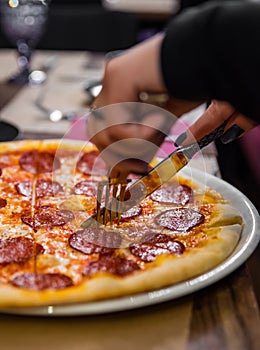 Woman hands with knife and fork cutting pepperoni pizza on table