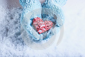 Woman hands in light teal knitted mittens are holding beautiful entwined vintage red heart on snow. Love, St. Valentine concept.