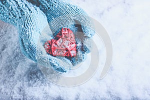 Woman hands in light teal knitted mittens are holding beautiful entwined vintage red heart on snow. Love, St. Valentine concept.