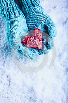 Woman hands in light teal knitted mittens are holding beautiful entwined vintage red heart in a snow. St. Valentine concept.