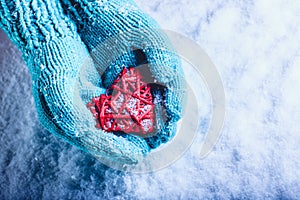 Woman hands in light teal knitted mittens are holding beautiful entwined vintage red heart in a snow. St. Valentine concept.