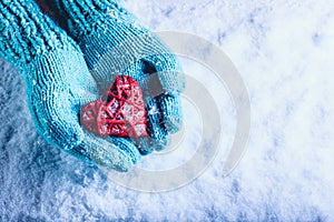 Woman hands in light teal knitted mittens are holding beautiful entwined vintage red heart in a snow. St. Valentine concept.