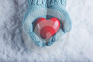 Woman hands in light teal knitted mittens are holding beautiful glossy red heart in snow background. Love, St. Valentine concept