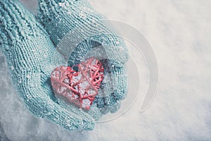 Woman hands in light teal knitted mittens are holding beautiful glossy red heart on snow background. Love, St. Valentine concept