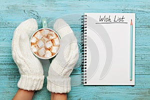 Woman hands in mittens hold cup of hot cocoa or chocolate with marshmallow and notebook with wish list on turquoise vintage