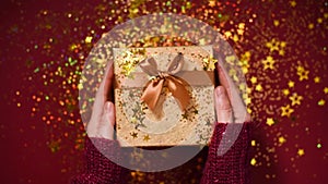 Woman hands opening gift box. Sparkling gold stars, glitter confetti over present. Unpacking gift on red background