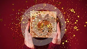 Woman hands opening gift box. Sparkling gold stars, glitter confetti over present. Unpacking gift on red background
