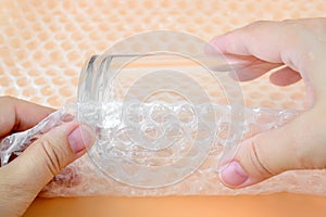 Woman hands packaging a glass for water with white transparent bubble wrap on a yellow background. Material for packing fragile