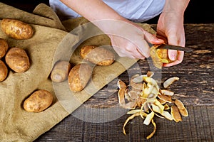 Woman hands peel potato, peelings on wooden cutting board. Three clean potatoes on plate.