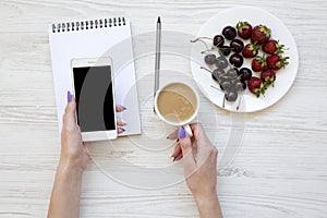 Woman hands with smartphone, latte, notebook, strawberries and cherries on white wooden background, top view.