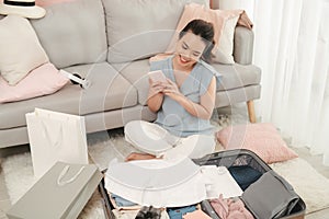 Woman hands using smartphone write checklist to prepare accessories for new journey, packing clothes in suitcase bag
