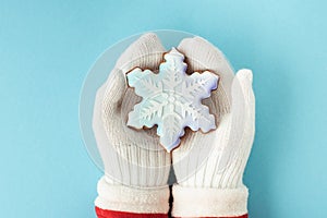 Woman hands wearing white mittens and holding snowflake shaped gingerbread cookie with festive icing on the blue pastel background