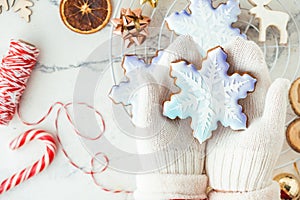 Woman hands wearing white mittens and holding snowflake shaped gingerbread cookie with festive icing on the decorated Christmas