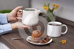 Woman hands with white kettle tea cup and choccolate cookie. Selective focus. Kitchen teatime. Time for rest and home