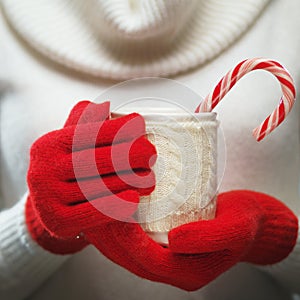 Woman hands in woolen red gloves holding a cozy mug with hot cocoa, tea or coffee and a candy cane.