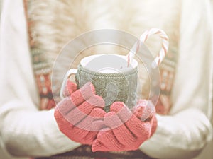 Woman hands in woolen red gloves holding a cozy mug with hot cocoa, tea or coffee and a candy cane. Winter and Christmas concept.