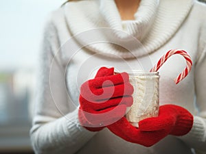 Woman hands in woolen red gloves holding a cozy mug with hot cocoa, tea or coffee and a candy cane. Winter and Christmas concept.