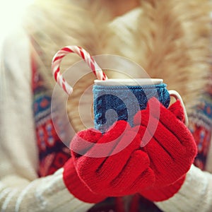 Woman hands in woolen red gloves holding cozy mug with hot cocoa, tea or coffee and candy cane. Winter and Christmas time concept.