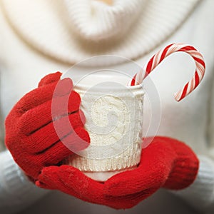 Woman hands in woolen red gloves holding a cozy mug with hot cocoa, tea or coffee and a candy cane.