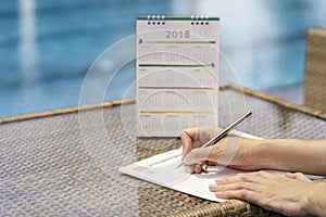 Woman hands writing plan on notebook, planning agenda and schedule using calendar event planner on desk near swimming pool