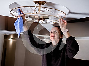 a woman happily wipes the ceiling chandelier from dust on a cozy evening. Apartment cleaning process