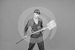 woman hard worker in protective helmet and boilersuit hold shovel on orange background, labour day
