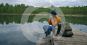 A woman with a hat and a backpack sits on a wooden pier of a forest lake in the mountains and drinks hot drinks tea or