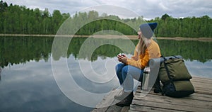 A woman with a hat and a backpack sits on a wooden pier of a forest lake in the mountains and drinks hot drinks tea or