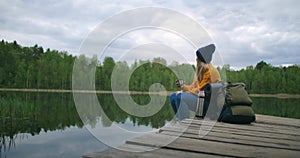A woman with a hat and a backpack sits on a wooden pier of a forest lake in the mountains and drinks hot drinks tea or