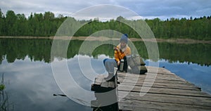 A woman with a hat and a backpack sits on a wooden pier of a forest lake in the mountains and drinks hot drinks tea or