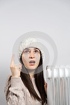 A woman in a hat getting warm near the oil heater and looks and points up with her finger.