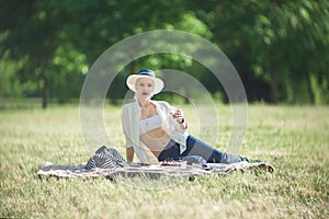 A woman in a hat is holding a glass of red wine in her hand at a picnic. Lady sitting on the grass with a blanket